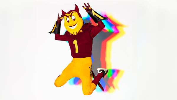Sparky the Sun Devil with multi-colored shadows jumping in front of a white background.