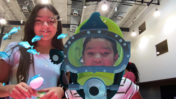 Two young students smiling at the camera, one is wearing an underwater breathing helmet while fish swim around his head.
